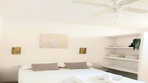 A neatly made bed with white linens and two pillows, a ceiling fan, and a wall-mounted shelf with baskets and a plant.