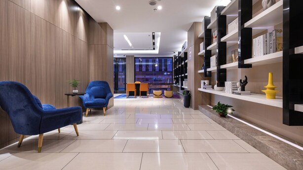 A modern hotel lobby with a wooden wall, blue chairs, a bookshelf, and a seating area with a table and chairs.