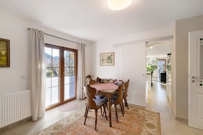 A dining area with a table and chairs, a radiator, a window with curtains, and a door leading to another room.