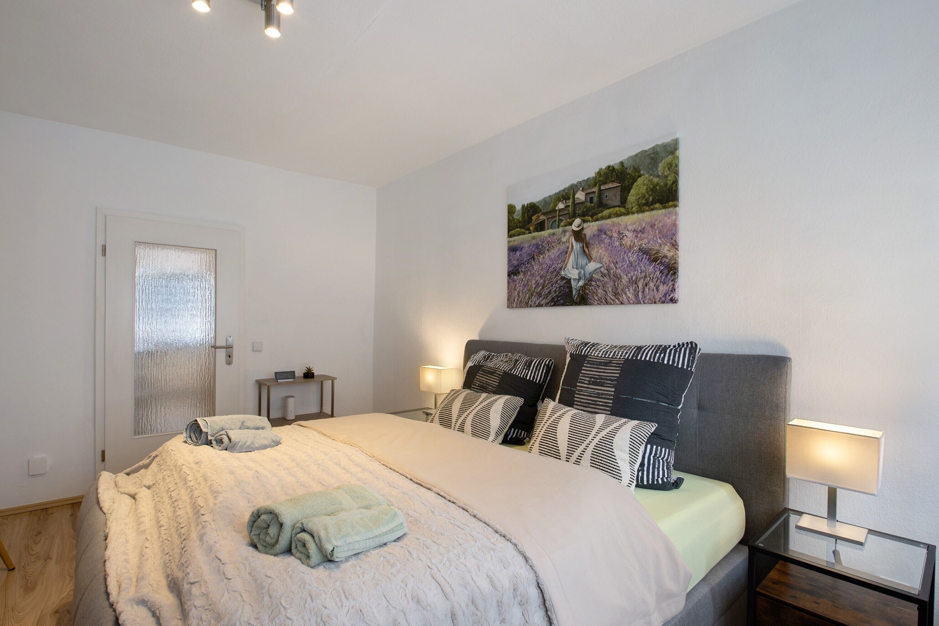 Un lit bien fait, avec une couverture et des serviettes pliées, une table de chevet avec une lampe et un tableau représentant un paysage accroché au mur.