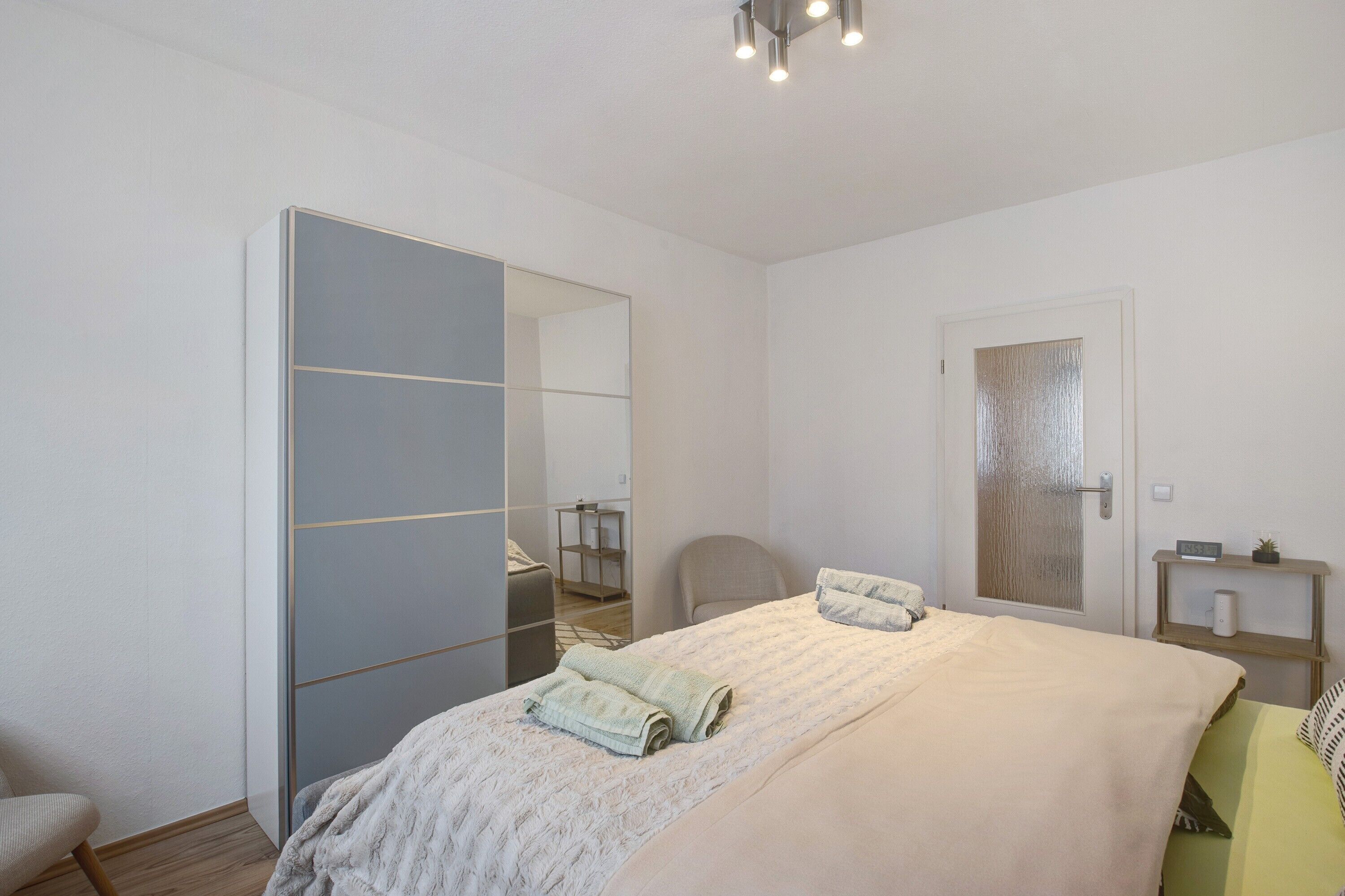 Ein Schlafzimmer mit Bett, Stuhl, Kleiderschrank, einer Tür und einem Regal mit einer Pflanze.