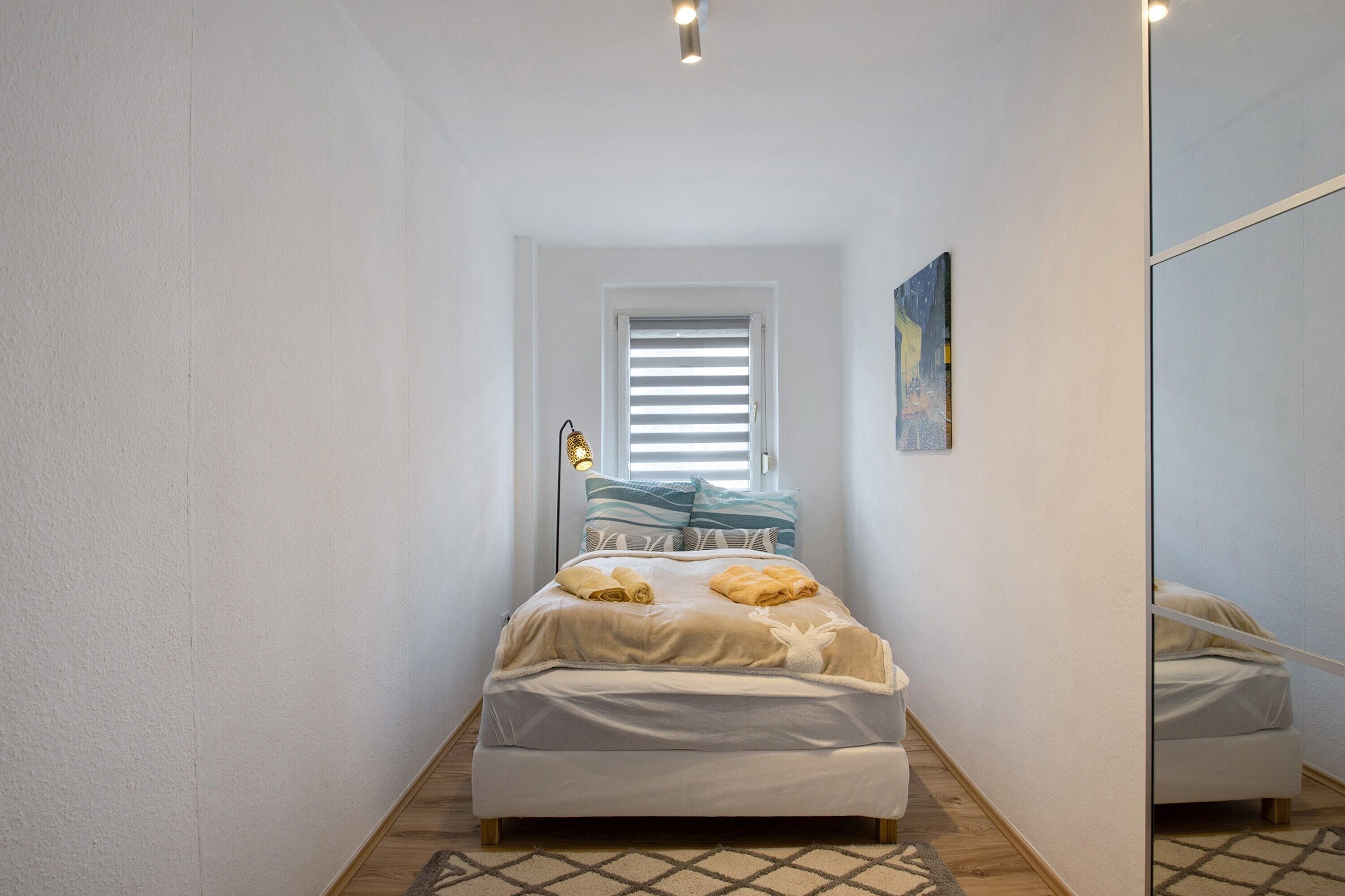 Une chambre à coucher étroite et bien éclairée, avec un lit, une fenêtre avec des stores, une lampe de chevet et un tableau au mur.