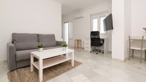 A modern living room with a grey sofa, a white coffee table, and a small plant. There is a TV mounted on the wall, a desk with a chair, and a window with blinds.