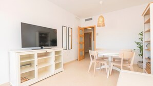 A modern living room with a flat-screen TV, a white entertainment unit, a dining table with chairs, and a bookshelf.