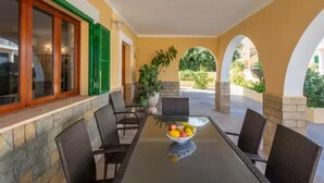 A covered outdoor dining area with a glass table, wicker chairs, and a bowl of fruit.