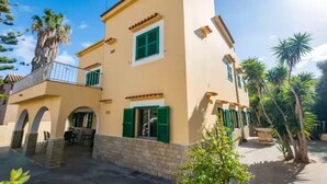 A two-story building with a balcony, green shutters, and a patio area with chairs.