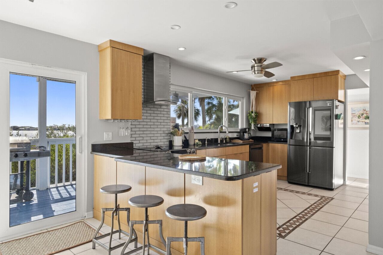 A modern kitchen with a large island, bar stools, a stainless steel refrigerator, and a view of the outdoors.