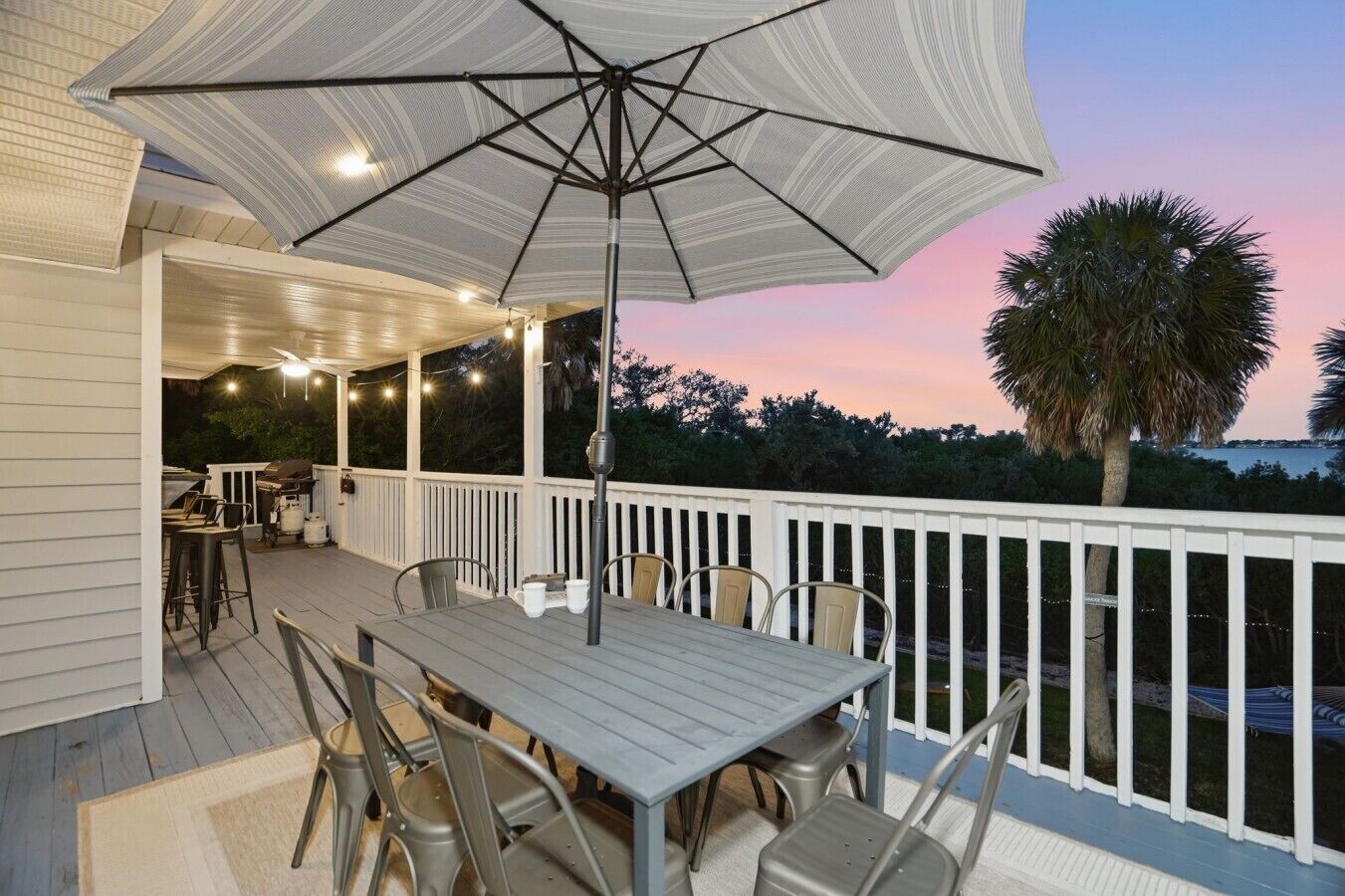 An outdoor deck with a table and chairs, a large umbrella, and a view of the ocean at dusk.