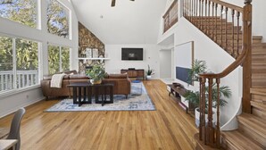 A spacious living room with a wooden staircase, a ceiling fan, and a large window overlooking trees. - Mangrove Manor | Waterfront + Backyard Oasis (Seminole)