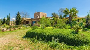 Un bâtiment traditionnel en pierre, doté d’une tour, entouré d’un jardin bien entretenu où poussent différentes espèces de plantes, le tout sous un ciel dégagé.