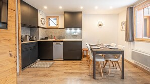 A compact kitchen with dark cabinets, a dishwasher, and a dining area with a table and chairs. - Smartstay Cristal - Appartement au pied des pistes (Méribel)