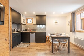 A compact kitchen with dark cabinets, a dishwasher, and a dining area with a table and chairs. - Smartstay Cristal - Appartement au pied des pistes (Méribel)