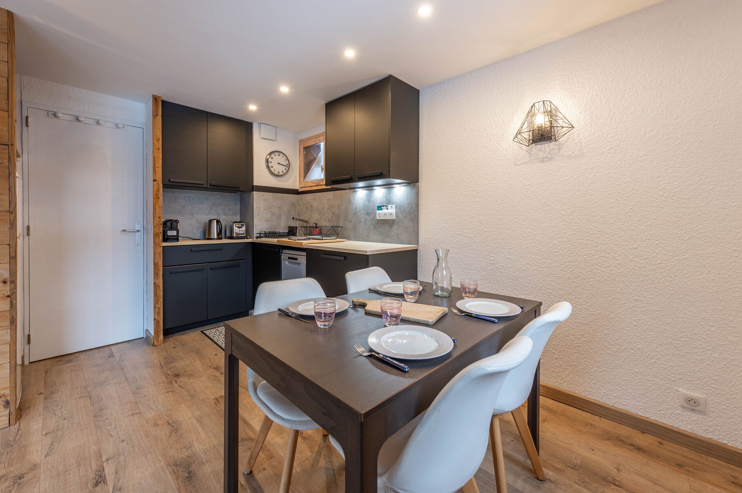 A modern kitchen with dark cabinets, a dining table set for four, and a white wall with a geometric light fixture.