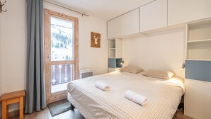 A bedroom with a large bed, a window with curtains, a wooden bench, and built-in shelving. - Smartstay Cristal - Appartement au pied des pistes (Méribel)