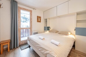 A bedroom with a large bed, a window with curtains, a wooden bench, and built-in shelving. - Smartstay Cristal - Appartement au pied des pistes (Méribel)