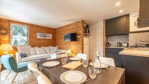 A modern living room with a wooden wall, a sofa, a dining table set for four, and a kitchen area with a microwave and toaster. - Smartstay Cristal - Appartement au pied des pistes (Méribel)