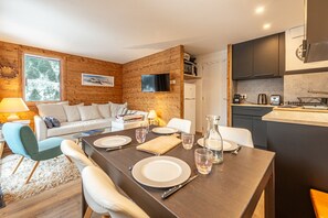 A modern living room with a wooden wall, a sofa, a dining table set for four, and a kitchen area with a microwave and toaster. - Smartstay Cristal - Appartement au pied des pistes (Méribel)