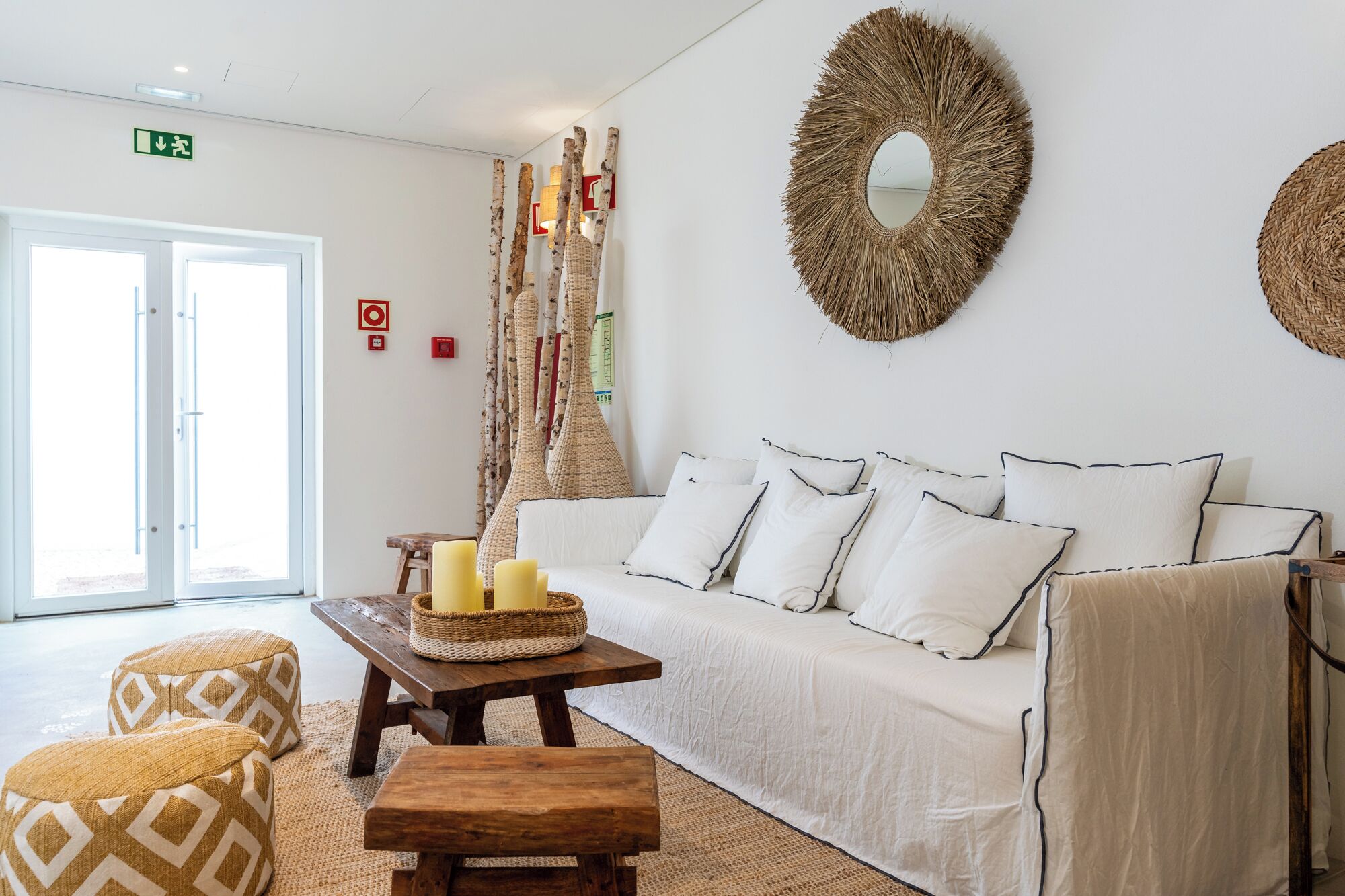 A modern hotel room with a white sofa, patterned cushions, a wooden bench, and a woven wall hanging.