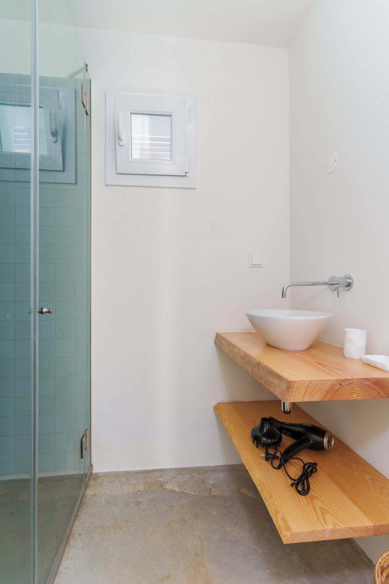 A bathroom with a glass shower enclosure, a white sink with a wooden countertop, and a hairdryer on a wooden shelf.