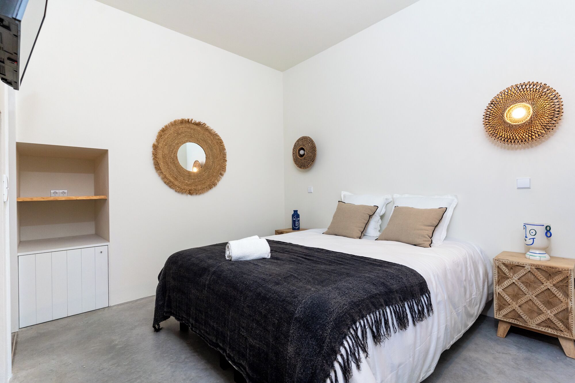 A bedroom with a bed, a bedside table, a shelf with a mirror, and a wall-mounted decorative item.