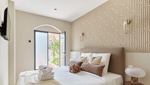 A modern hotel room with a large bed, a flat-screen TV, a bedside lamp, and a view of the outdoors through a glass door.