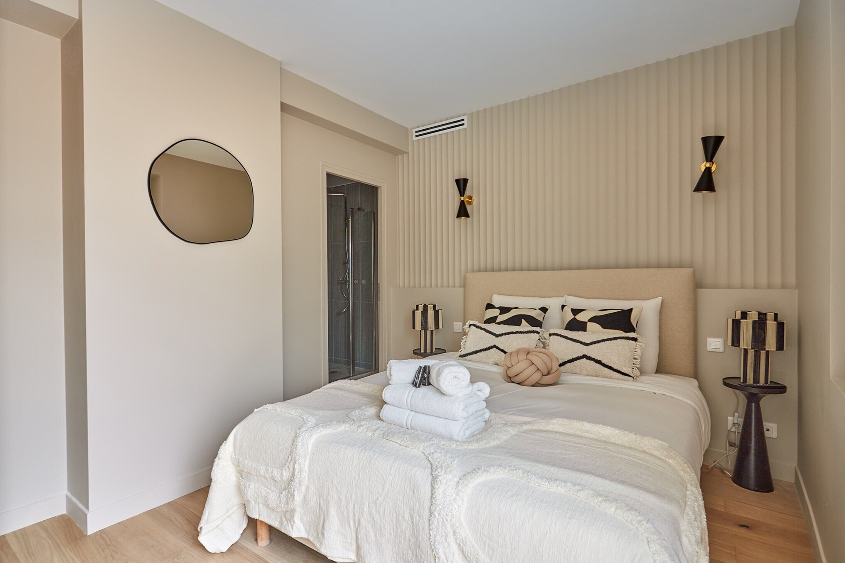 Une chambre à coucher avec un grand lit, un miroir rond, deux appliques murales et une table de chevet avec une lampe.