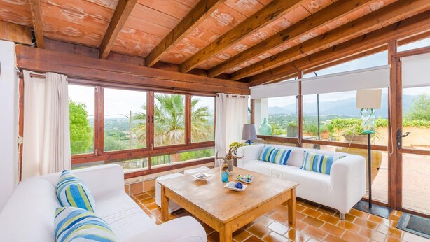 Ein geräumiger Raum mit Holzboden, großen Fenstern mit Blick auf Palmen und Berge, einem weißen Sofa mit gestreiften Kissen und einem Holztisch mit Vase und Tellern.
