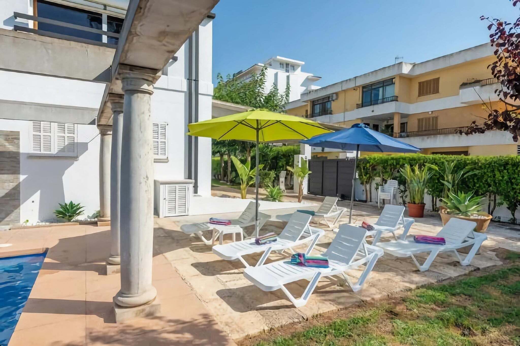 A poolside area with white lounge chairs and umbrellas, a clear sky, and modern buildings in the background.