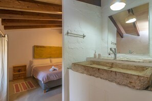 A bedroom with a bed, bedside table, and a wooden headboard. There is a bathroom with a sink, mirror, and towel rack.