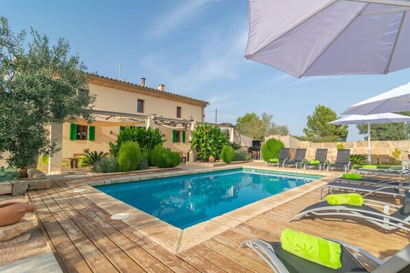 A swimming pool with lounge chairs and umbrellas, surrounded by greenery and a building with a tiled roof.