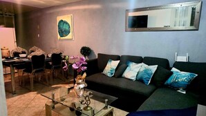Sala de estar con sofá, mesa de centro y sillas. Hay un cuadro en la pared y una ventana con persianas.