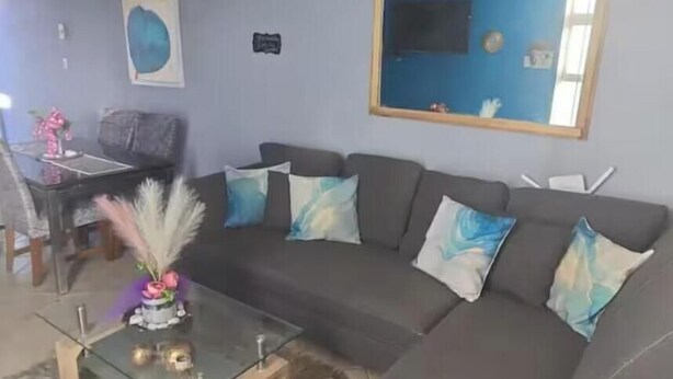 Una sala de estar con un sofá seccional, una mesa de centro de vidrio, una alfombra mullida, un cuadro en la pared y una zona de comedor al fondo.