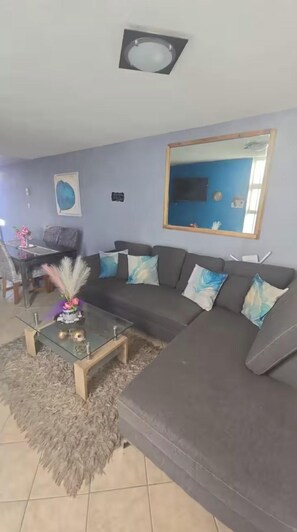 Un salon avec un canapé d’angle, une table basse en verre, un tapis moelleux, un tableau au mur et, au fond, un coin repas.
