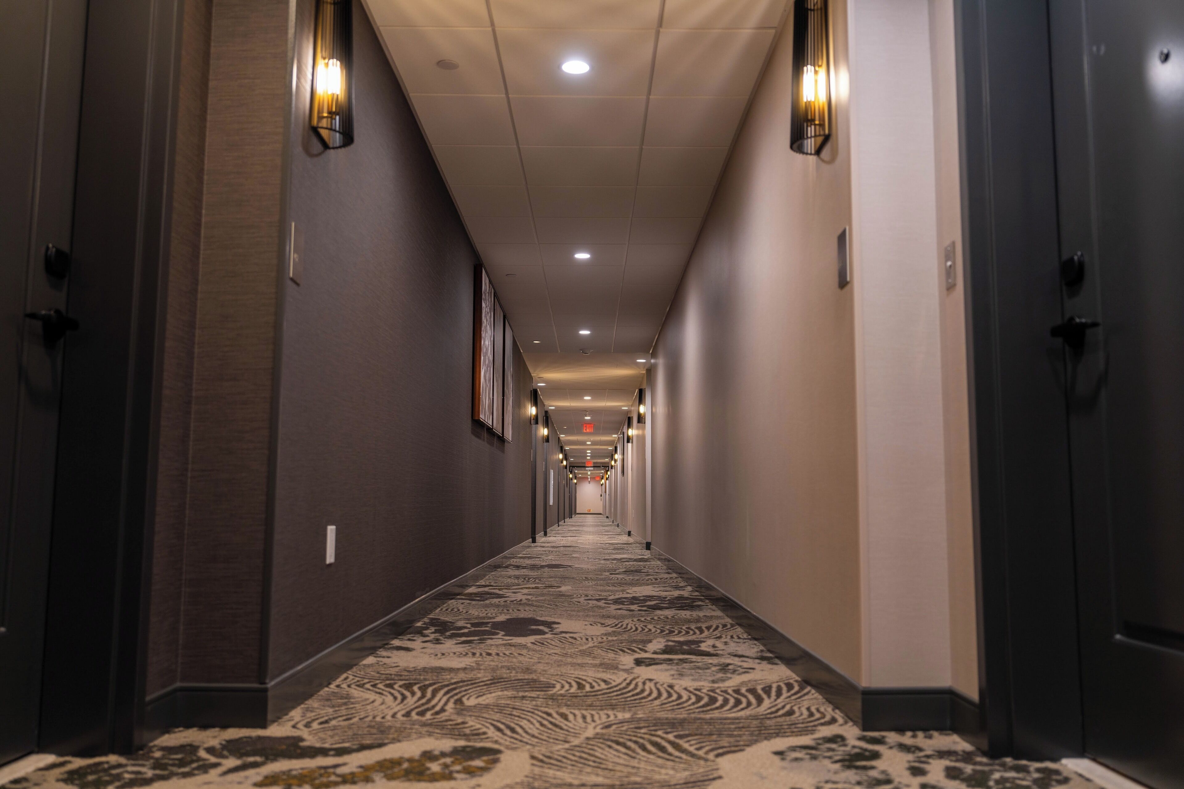 A long, narrow hotel corridor with patterned carpet, recessed lighting, and doors on both sides.