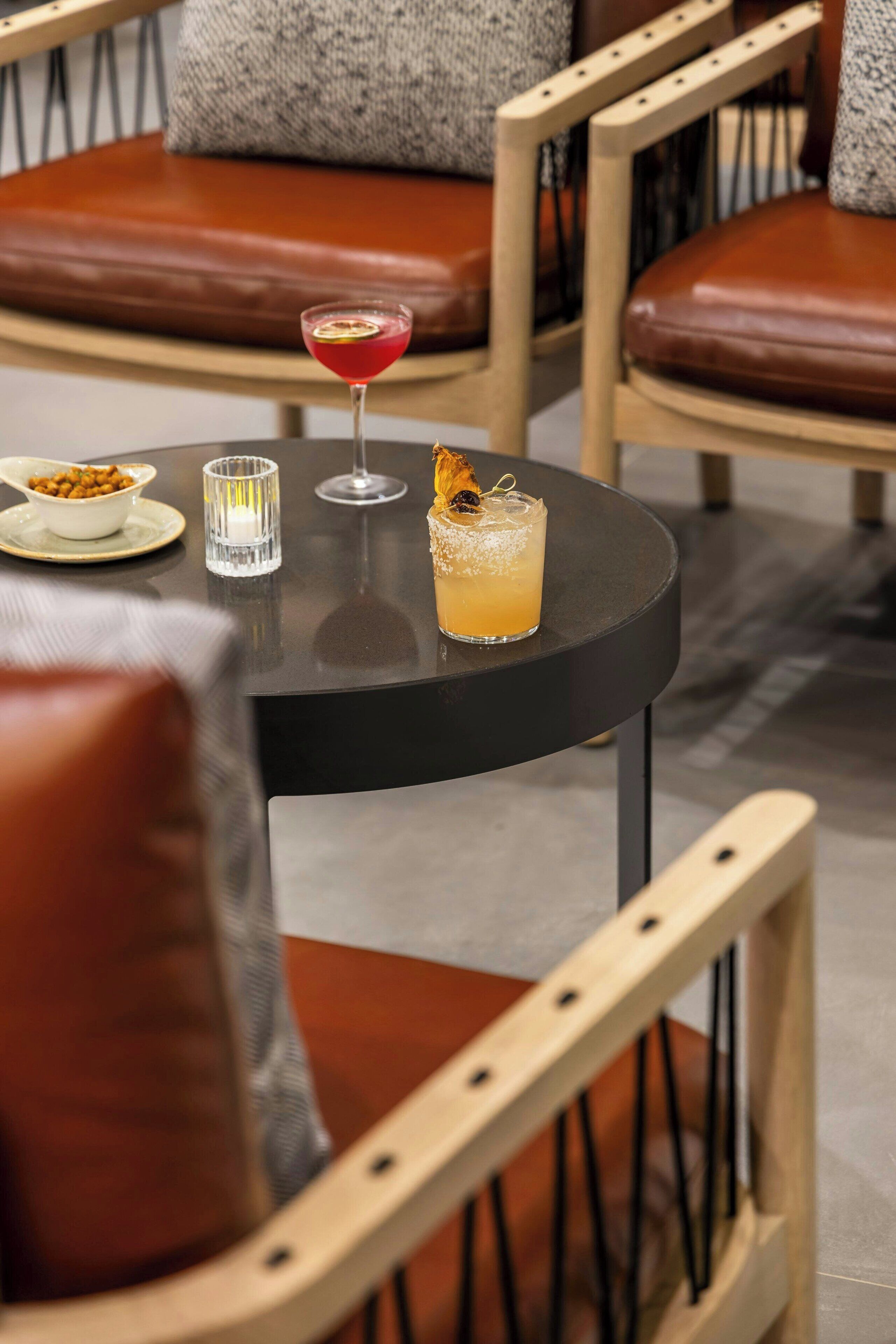 A round table with a glass of red cocktail, a glass of orange juice, and a candle. There are brown leather chairs around the table.