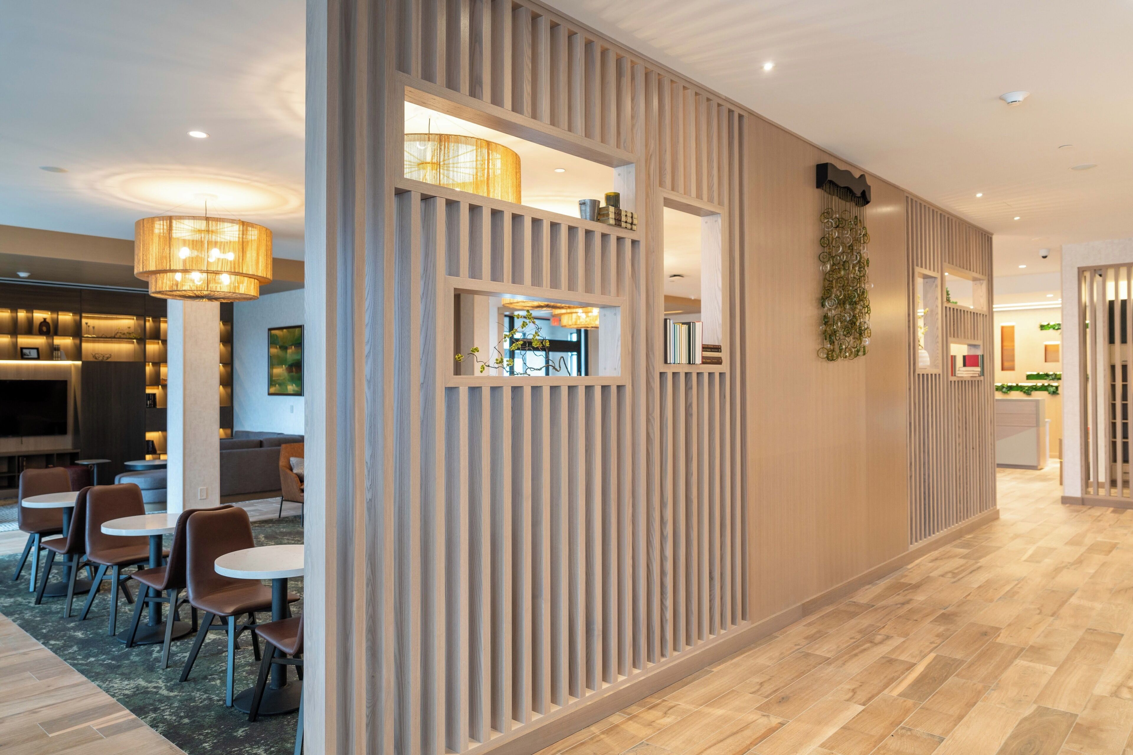 A modern hotel lobby with a wooden reception desk, seating area with round tables and chairs, and a large chandelier.