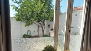 Ein Holztisch mit kariertem Tuch, ein Blumentopf, ein weißer Stuhl und Blick auf einen Garten mit Baum und weißer Mauer.