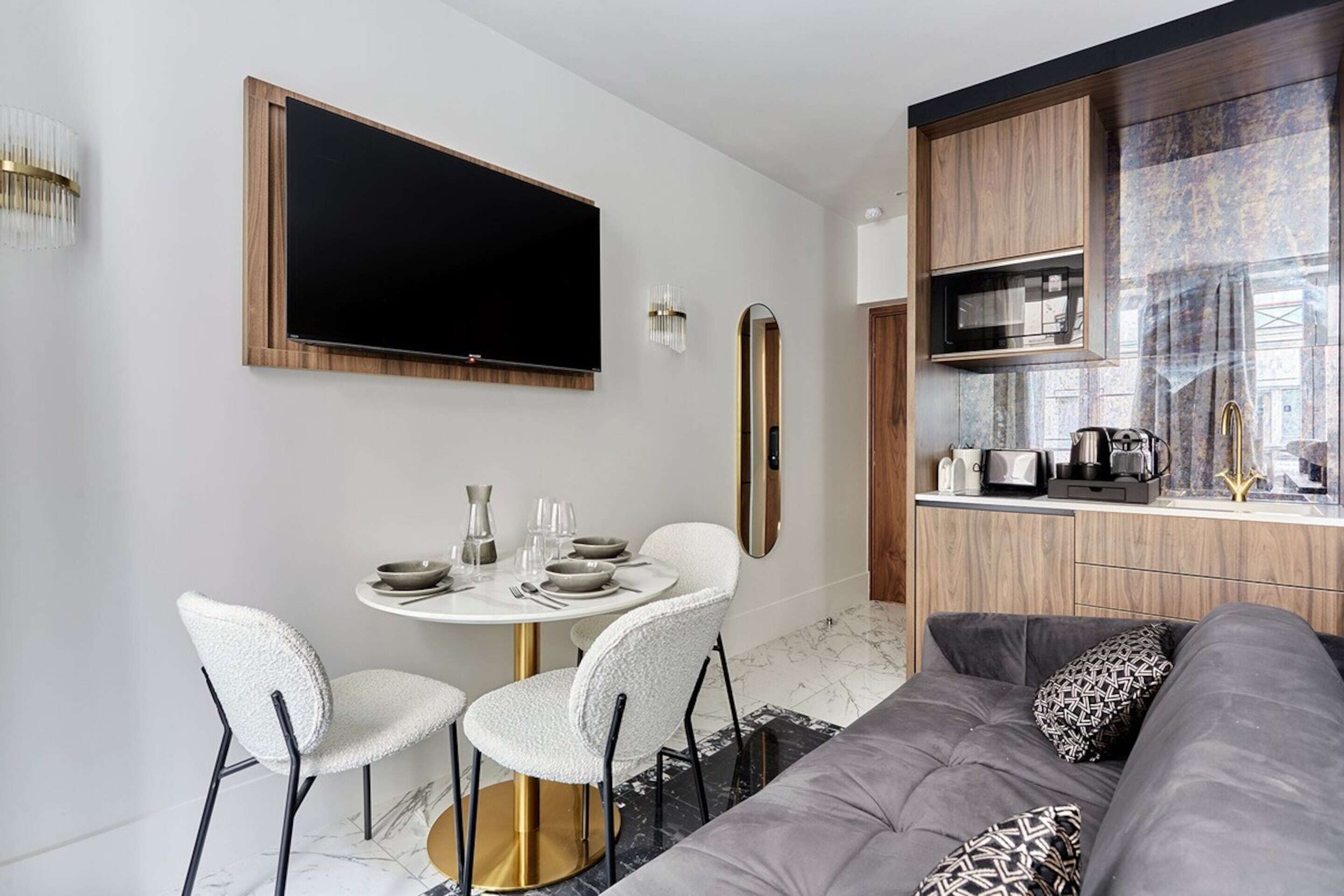 A modern living room with a grey sofa, a round dining table with white chairs, a flat-screen TV mounted on the wall, and a kitchen area with wooden cabinets and a microwave.