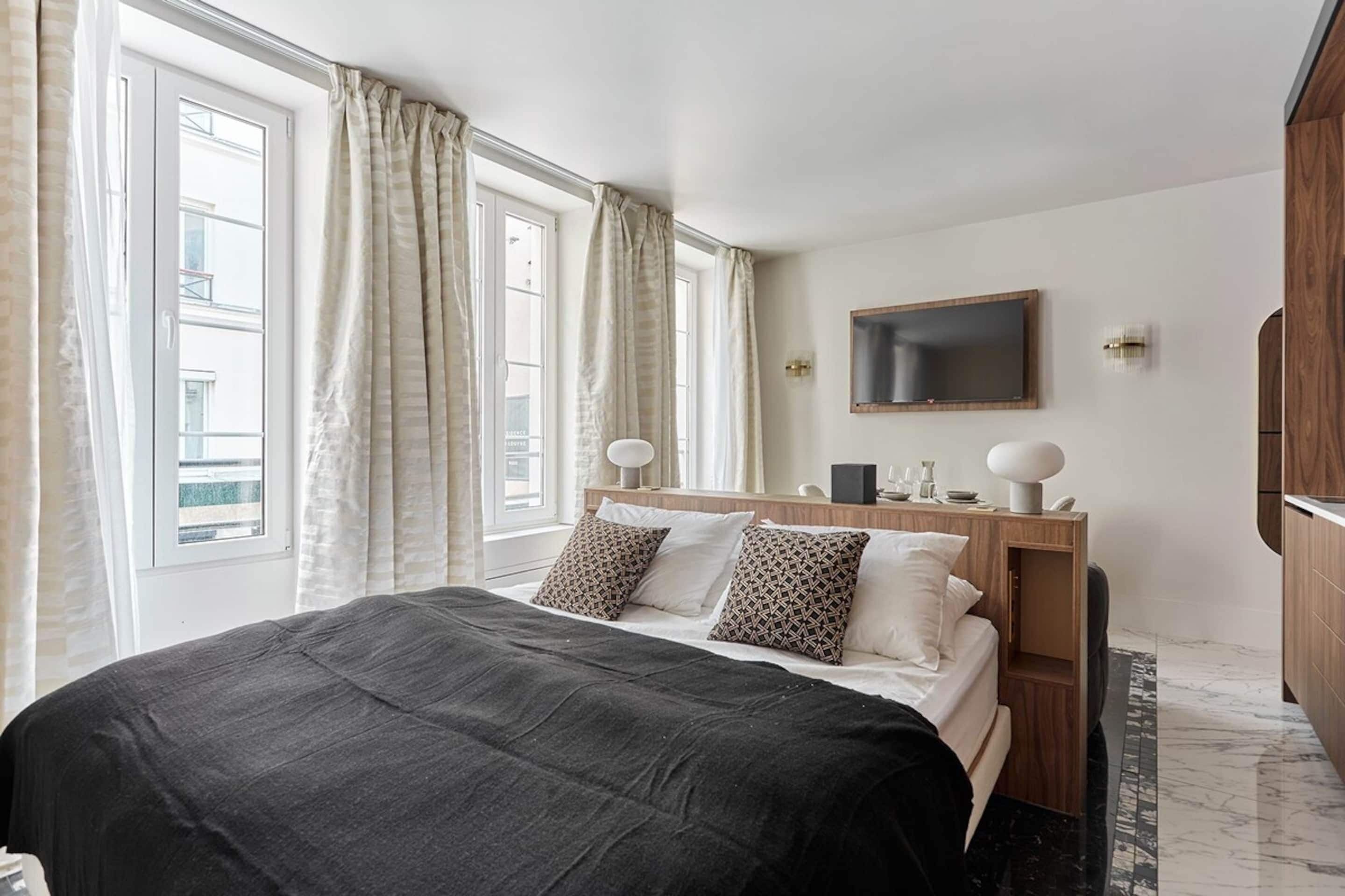 Quarto de hotel com cama, cortinas, televisão e mesa de cabeceira de madeira.