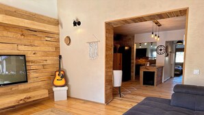 A modern living room with a wooden wall, a TV, a guitar, a couch, and a kitchen in the background. - Alba | Air-conditioned, Wi-Fi, PMR, beach 20 metres away (Borgo)
