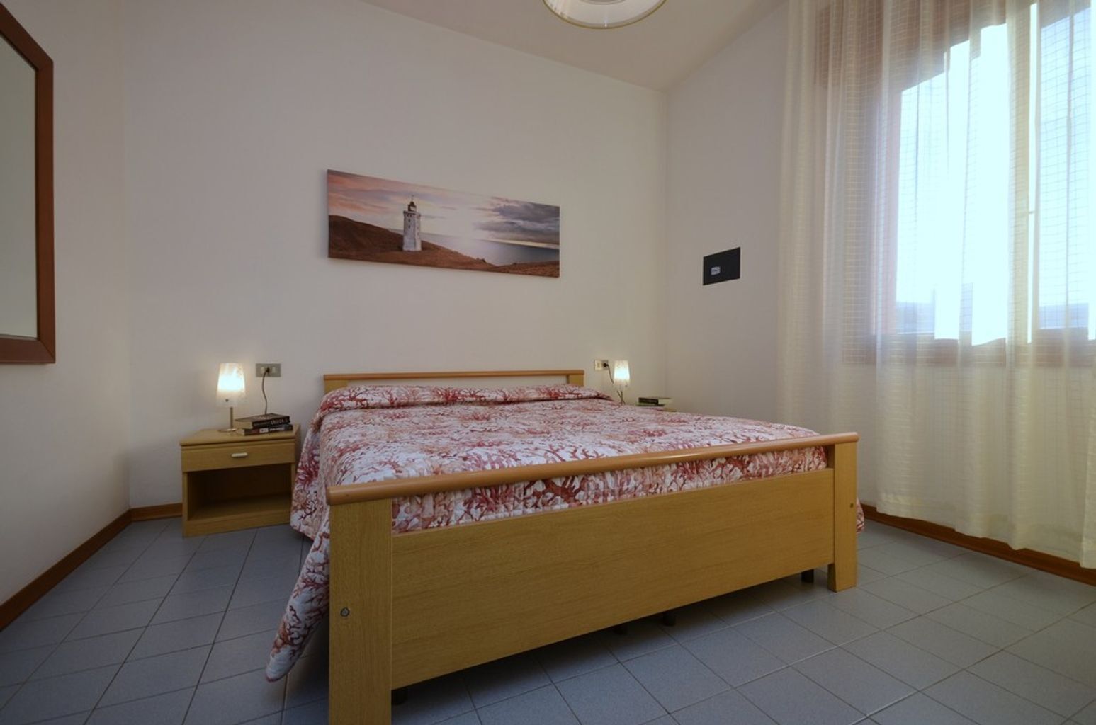 A bedroom with a wooden bed, two bedside tables with lamps, a painting on the wall, and a window with curtains.