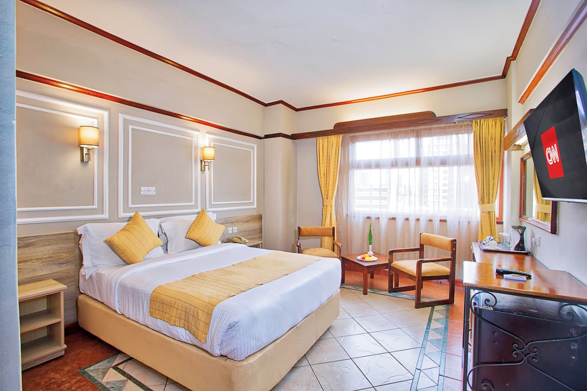 A hotel room with a large bed, bedside tables, a desk with a chair, a TV, and a window with curtains.