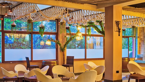 Un espace repas avec des chaises et des tables en osier, de grandes fenêtres donnant sur la verdure et des décorations murales en macramé.