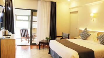 A hotel room with a bed, a desk, a chair, a table with a plant, and a view of the outside.