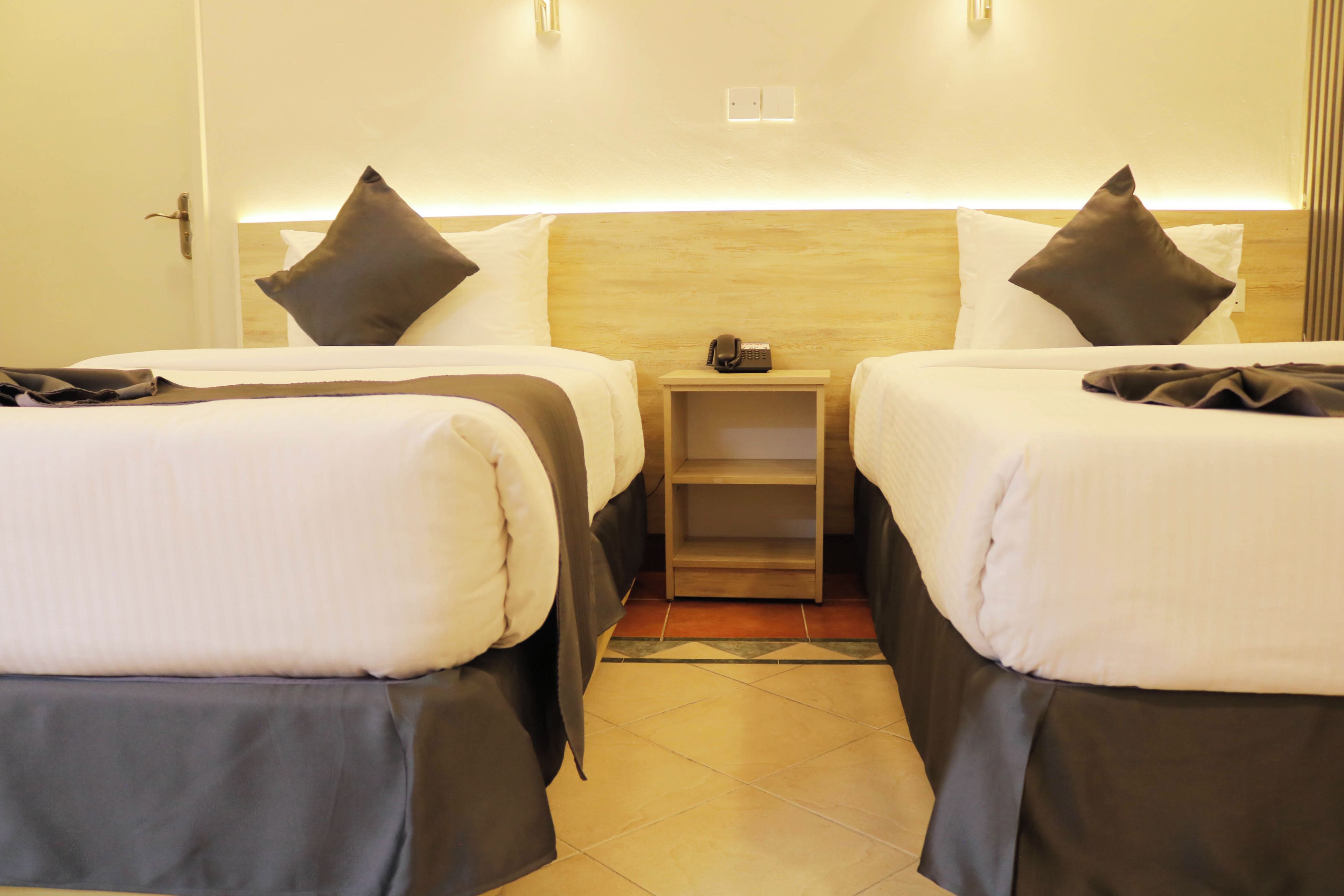 A hotel room with two single beds, each with a pillow and a blanket, a bedside table with a phone, and a small shelf.