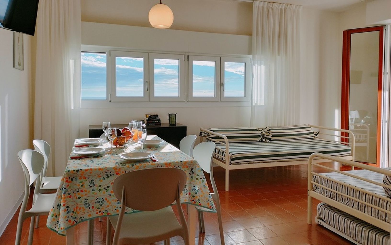 Ein Zimmer mit Esstisch, gedecktem für vier Personen, einem Bett mit gestreifter Bettwäsche und Meerblick durch große Fenster.