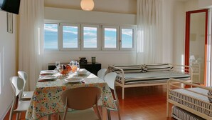 Ein Zimmer mit Esstisch, gedecktem für vier Personen, einem Bett mit gestreifter Bettwäsche und Meerblick durch große Fenster.