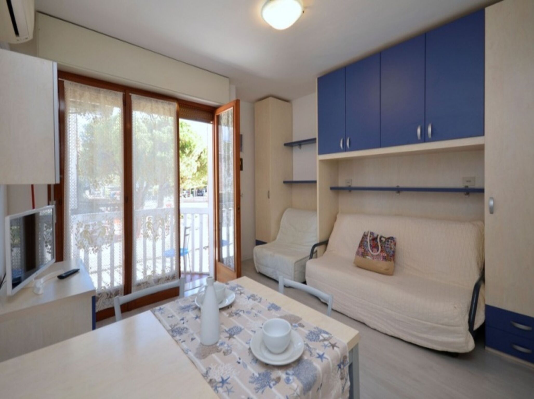 Quarto com mesa posta para dois, sofá, televisão e varanda com vista.