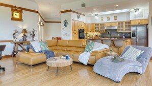 A modern living room with a sofa, armchair, and coffee table. A kitchen with wooden cabinets and appliances is visible in the background. - Scenic Views with Pool Table & Hot Tub | Cathedral View by AvantStay (Sedona)