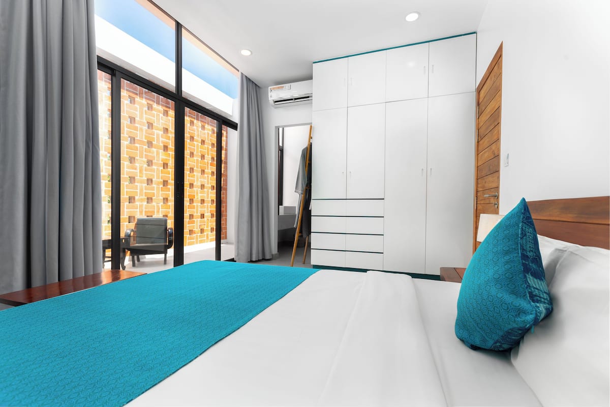 A modern hotel room with a large bed, a view of a brick building, a wardrobe, and a desk with a chair.